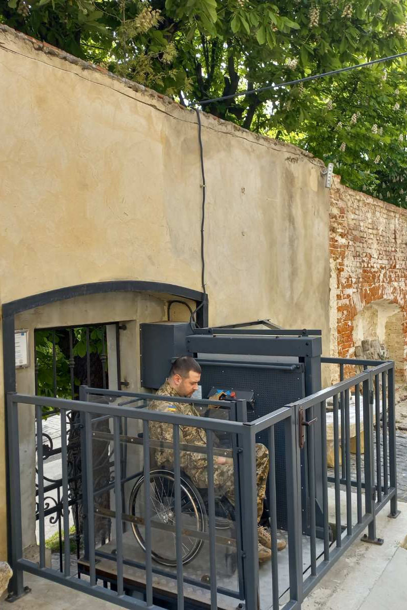 Воїн в кріслі колісному піднмається гідравлічною платформою на подвірʼя музею