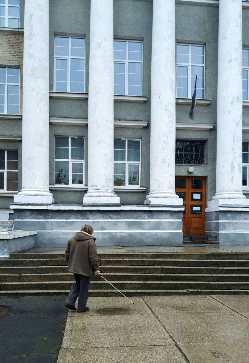 Людина з порушенням зору підходить до сходів перед фасадом музею